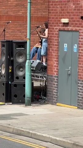 A person wearing a dark halter top and denim shorts is playing a guitar and singing into a microphone. They are standing on a stage setup next to a brick building with a grey metal door. To the left of the stage are several large black speakers. A green metal pole is positioned in front of the stage and speakers. Colorful pennant banners are strung above the brick building. Another person is seated on the stage behind the singer. The scene appears to be outdoors on a paved surface, possibly a street.