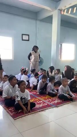 The video shows a room with light blue walls, a white ceiling, and light-colored tiled floors. A light blue pillar is visible in the center of the room. A large, patterned red rug covers the floor in the foreground. On the rug, a group of young children, appearing to be elementary school age, are sitting on the floor. They are dressed in light-colored shirts and dark pants or shorts. Some of the children are wearing headbands with decorations, such as bunny ears. Two adult women are also present in the room. One woman, wearing a light-colored vest over a shirt and pants, stands on the left side of the room, facing the children. Another woman, wearing a headscarf and a dark top, is seated on the right side of the room, also facing the children. A banner with text that appears to say "JESS EDUCATION" is visible on the back wall, along with some decorative flags hanging from the ceiling. There are windows on the back wall, and a fan is mounted on the wall. The children appear to be attentive and engaged with the standing woman.
