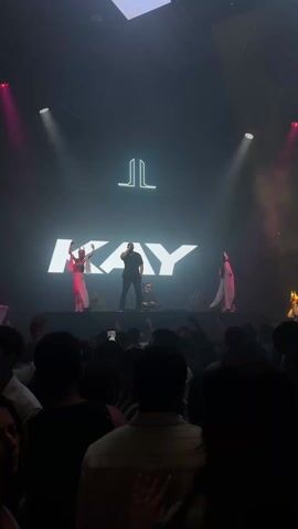 A person onstage, illuminated by bright lights, is performing in front of a crowd.  Behind them, a large screen displays "KAY" in white letters. Two dancers in matching dresses stand on either side of the performer. A DJ can be seen behind the performer. The crowd is primarily composed of people standing and holding up their phones to record the performance. The venue appears to be a nightclub or concert hall, with a dark ceiling and spotlights.