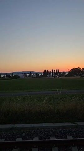 The video shows a landscape at sunset. The sky transitions from a clear blue at the top to orange and pink near the horizon. In the distance, there is a body of water, likely a lake, with hills or mountains on the other side. A line of trees stands against the sky in the mid-ground. There are some buildings visible on the right side of the frame, including a modern multi-story building. In the foreground, there is a grassy field and a train track. The lighting suggests it is late evening. There are no people or animals visible in the video.