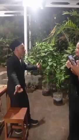 A man in a black suit and cap is standing and gesturing with his hands to a woman holding a microphone. The woman is also wearing black. They appear to be outdoors at night, with plants and a roof overhead. The man is standing next to a wooden stool, and there is a table to his left.