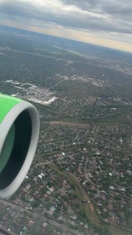 The video shows an aerial view from an airplane window. In the foreground is the airplane's engine cowling, painted white with a green rim. Below, the ground is visible, showing a densely populated suburban area with houses, trees, roads, and some commercial developments. A set of train tracks runs through the landscape. The sky above is partly cloudy with patches of sunlight breaking through.