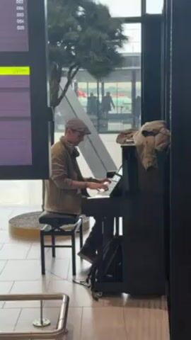 A man wearing a cap and jacket is playing a piano at an airport. He is seated on a black stool, facing the piano. A black backpack rests on the floor beside the piano. To the left of the piano is a flight information display showing departure gates and times. Behind the man, through a large window, people can be seen outside. The floor is tiled, and there is a circular rug in the foreground. A coat is draped on top of the piano.