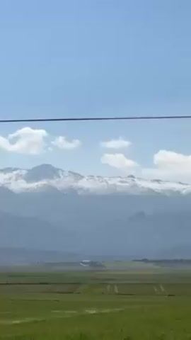 The video shows a landscape with a bright green field in the foreground. Beyond the field, there are hazy mountains, the peaks of which are covered in snow. The sky is blue with a few fluffy white clouds. An electrical wire is visible across the top of the frame, and power poles can be seen throughout. The perspective is from a moving vehicle. There is a sound of wind.