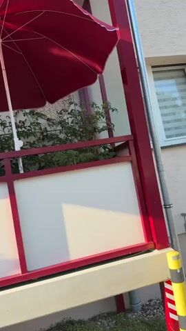 This media file presents a close-up, slightly tilted view of a balcony on a sunny day. The primary subjects are a large, open, dark red patio umbrella and a red railing that encloses the balcony. Behind the railing, a window with white frames is visible, reflecting the sky and surrounding trees. Green plants, appearing to be tomato plants, are growing in a planter box behind the railing, and a cluster of vibrant red flowers, possibly geraniums, is also present.

The scene suggests a leisurely outdoor setting, likely a residential balcony. The time of day appears to be late afternoon or early evening, indicated by the warm, angled sunlight casting distinct shadows on the white panels of the balcony railing and the ground below. The weather is clear and bright. The location context of Rathenow, Germany, places this scene in a European urban or suburban environment.

There are no people visible in the frame. The overall mood is peaceful and serene, enhanced by the natural elements and the gentle sunlight. Objects visible include the umbrella, railing, window, plants, flowers, a small decorative wicker item on the railing, and what appears to be a drainpipe at the bottom of the frame. The white panels of the railing are illuminated by the sun, contrasting with the red frame. The reflection in the window adds depth to the scene, hinting at the wider environment outside the balcony.