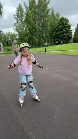 A young girl with blonde hair wearing a pink t-shirt, ripped denim jeans, a polka-dot helmet, elbow pads, and knee pads is rollerblading on an asphalt path. She is looking down and smiling. To her left and in the background, an older man in a light-colored shirt and blue jeans is also on the path, walking away from the camera. In the further background, there are trees and a grassy area. The camera moves closer to the girl, then pans down to show a blurry image of a small, fluffy dog walking away from the camera on the asphalt.