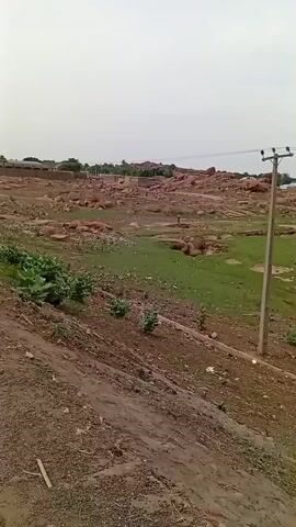 The video opens with a wide, elevated shot of a dry, uneven landscape featuring scattered green patches and numerous piles of reddish-brown material, possibly rocks or construction debris. Distant buildings are visible on the horizon under an overcast sky. The camera then pans left, revealing a dirt road with vehicular traffic, including cars and motorcycles, moving in both directions. Streetlights line the road. The camera continues to pan, showing more of the dusty terrain, green shrubbery, and a utility pole with overhead power lines. In the background, a small settlement with buildings and trees can be seen, bordering a flatter, grassy area. A fence and a few indistinct human figures are also visible in the distance.