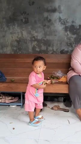 A young child wearing a pink outfit and blue sandals stands and walks. The child is holding green fruits. The child walks towards a broom. The child is near a wooden bench and a wall. There is a bottle of water and more green fruits on the bench. There are also shoes and other items on the floor. A person's leg is visible in the lower left corner of the frame.