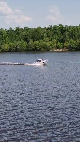 The video shows a white speedboat moving across a dark body of water, likely a river or lake, on a sunny day. The boat is moving from right to left, leaving a white wake behind it. The boat has a small cabin and an antenna on top. The water is calm, with small ripples. On the far bank of the water, there is a dense line of green trees. The sky is light blue with a few small white clouds. The scene is captured with a slight movement of the camera, though the focus remains centered on the boat.