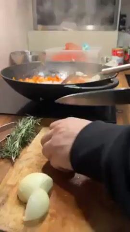 A man in a black long-sleeved shirt stands at a wooden counter, chopping an onion on a wooden cutting board. To the left of the onion is a sprig of rosemary and a pair of tongs. On a stovetop in the background, a large, dark-colored wok is set to cook. Inside the wok are diced carrots and onions, as well as a metal spatula being used to stir the ingredients. Steam rises from the wok. Behind the wok are additional cooking utensils, a steel bowl, and a red container that appears to contain whole tomatoes. The kitchen has white walls and cabinets.