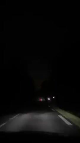 A vehicle travels on a dark road at night, illuminated by its headlights. Trees line both sides of the road. The taillights of another vehicle can be seen in the distance ahead. A triangular warning sign is visible on the right side of the road.