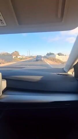 The media file shows a first-person perspective from inside a car, looking through the windshield. The car is driving on a paved road in Ahrara, Morocco. The scene is set during the daytime, under a clear blue sky with a few wispy clouds. The sun is shining, casting a warm light on the landscape.

In front of the car, another dark-colored car is traveling in the same direction. The road is bordered by dry, dusty terrain with sparse vegetation and some low-lying buildings or structures on either side. There are utility poles with wires visible along the road. The overall impression is a rural or semi-rural setting.

The interior of the car is visible in the foreground, with a dark dashboard and a section of the car's roof lining or sun visor. The camera is positioned to capture the road ahead and the passing scenery. There are no people clearly visible inside the car. The mood is peaceful and suggests a journey or travel. There is no visible text in the media file.