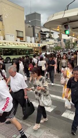 A medium shot shows a busy street crossing with many people walking across the street. The camera is positioned low, capturing the torsos and legs of the pedestrians. An older man in a white shirt and dark pants walks on the left with a cane and a black bag. A woman in a floral top and black pants walks beside him, carrying a light gray handbag. In the middle of the frame, a man in a black polo shirt and dark pants with a smartphone in hand walks while looking at the camera. In the background, a green bus and a red taxi are visible. People are moving in different directions, crossing the street. The weather appears overcast, and buildings line the street in the background.
