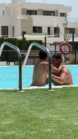 Two individuals are in a swimming pool. One individual is partially submerged, facing away from the camera and moving towards the pool steps. The other individual is also partially submerged, facing the camera, with their arms around the first individual. The scene takes place outdoors, with a building and artificial turf visible in the background.