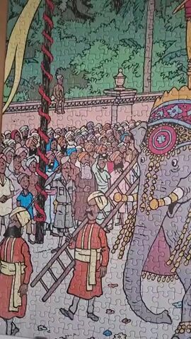 This is a close-up, static video of a jigsaw puzzle depicting a colorful parade scene. A large, decorated elephant is the central focus, adorned with intricate patterns and tassels. A golden palanquin sits atop the elephant, carrying several individuals in traditional attire. A crowd of people, also in traditional dress, surrounds the elephant, some carrying ladders. In the background, a wall and lush green foliage are visible. The puzzle appears to be in the process of being assembled or displayed.