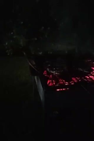 A grill with glowing red coals sits in darkness, with food cooking on the grate. Smoke rises from the grill. The background is dark, with indistinct shapes of trees and foliage. The scene appears to be outdoors at night.
