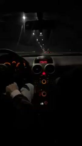 A driver navigates a car on a dark highway at night. The car’s dashboard glows dimly, illuminating the steering wheel, console buttons, and dashboard gauges. Outside the windshield, streetlights line the highway. Other cars are visible in the distance. The driver’s hands are on the steering wheel. Music plays in the background.