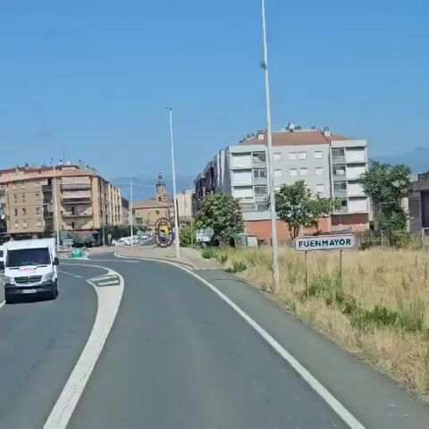 The video shows a view from a vehicle driving on a road. On the right side of the road, there is a sign that reads "100" indicating a speed limit. Further ahead, there is a circular sign with text around it, and a yellow speed limit sign that reads "40." The road curves to the left. In the background, there are buildings, a church tower, and trees. The sky is blue and clear.
