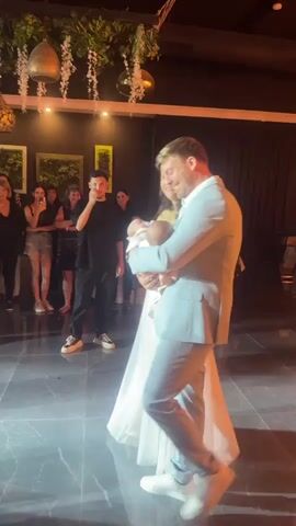 A wedding reception is in progress. The bride and groom are dancing on a polished floor. The groom, wearing a light-colored suit and white sneakers, holds a baby. The bride, in a white wedding dress, is facing him. Guests are watching from the background, many holding up their phones to record the moment. The room is decorated with hanging plants and ambient lighting. The overall atmosphere appears celebratory.