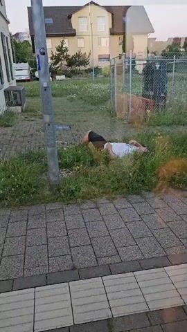 A person lies on their back in the grass next to a sidewalk. Their head is resting on the ground. Another person is partially visible, bent over them with their head down. A streetlamp is to the left of the frame. In the background is a building and a chain-link fence. The sky is light, suggesting it is daytime.