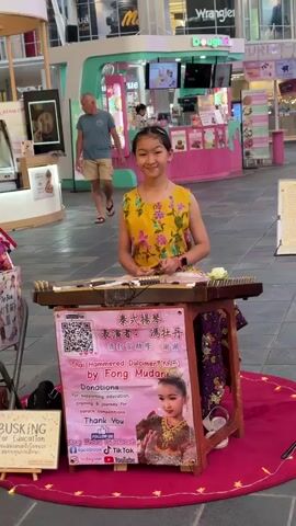 A young girl in a yellow floral top is playing a dulcimer on a street. She is sitting behind a wooden stand with a pink sign that reads "Thai Hammered Dulcimer (Khim) by Fong Mudan" and "Donations for supporting education, training & journey for karstic formations. Thank You". A QR code and social media icons are also visible on the sign. The girl is looking at the camera and smiling. In the background, a man in a blue shirt and shorts is walking past. Another man wearing a red hat and a black t-shirt is standing and looking at the girl. The environment appears to be a public space, possibly a mall or a marketplace, with shops and displays visible in the background. A red rug is laid out in front of the girl. A sign on the left side of the frame reads "BUSKING for Education".