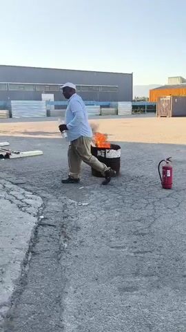 A man in a light blue shirt and tan pants walks to the right, away from a barrel on fire. The barrel is emitting black smoke and orange flames. A red fire extinguisher stands to the right of the barrel. The scene appears to be an outdoor industrial area with a large building in the background.