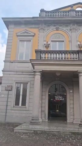 The video shows the exterior of a building with columns and arched windows. The building appears to be a theater, with signage that reads "TEATRO SOCIALE." There are trees and plants surrounding the building, and a car is parked in front. The sky is overcast. The video pans across the building, showing different architectural details and the surrounding environment.