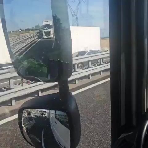 The media file shows a view from inside a vehicle, likely a truck, driving on a highway. The perspective is through the windshield and side mirrors. The main subject is an older, light-colored car with extensive modifications, including a luggage rack on the roof piled high with items. The car appears to be driving in the right lane of the highway, and the truck is in the lane to its left, observing it.

The highway has multiple lanes, separated by guardrails. In the distance, other vehicles, including trucks and vans, can be seen. The landscape outside the highway is flat and appears to be agricultural land, with fields of dry grass or crops. Tall power lines are visible in the distance. The weather appears to be clear and sunny, suggesting a daytime scene.

The car has visible text and graphics on its side, though much of it is blurry. A number "45" is clearly visible on the side of the car. Other markings and logos are present but unreadable. The car also has what looks like an aftermarket exhaust and lowered suspension.

The setting is a highway in Brugna, Italy, as indicated by the provided location context. The activity is simply traveling on the highway, with the focus on observing the unique car. There are no discernible emotions conveyed by the content. The overall impression is of a normal day of travel on an Italian highway, with a noteworthy vehicle being observed.