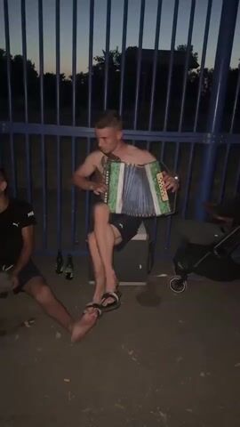 The media file captures a candid scene outdoors at dusk, likely in Saint-Orens-de-Gameville, France. The primary subject is a young, shirtless man with a tattoo on his chest, playing a green accordion. He is seated on what appears to be a cooler or storage box. He is wearing black shorts and flip-flops. His posture and facial expression suggest he is focused on his music.

To his left, another young man is visible, wearing a black t-shirt with a white logo that appears to be "Puma." He is sitting on the ground with his legs extended. Two green glass bottles, possibly beer bottles, are placed near him on the ground.

In the background, a blue metal fence runs horizontally across the frame, suggesting the location might be a park, sports facility, or a public area with such fencing. Beyond the fence, trees and a dark sky indicate it is either evening or night. A dark-colored stroller or pram is partially visible on the right side of the frame. The ground is dark and appears to be asphalt or a similar surface, with some wet patches.

The overall mood is relaxed and informal, possibly a social gathering or a moment of casual entertainment. The time of day is clearly evening, as indicated by the dim lighting and the deep blue of the sky. The weather appears to be calm. No specific text is clearly legible within the frame, other than the potential "Puma" logo on the t-shirt.