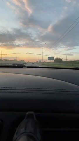 The view from the inside of a moving vehicle, possibly a car, shows a highway stretch at sunset. Water droplets are visible on the windshield. Several light poles line the highway. There's a road sign visible in the distance. The sky is filled with light pink-tinged clouds. The car's dashboard is textured and dark. There's a small, dark object fixed on the dashboard.