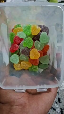 A close-up shot shows a clear plastic container filled with heart-shaped gummy candies in various colors, including red, green, yellow, orange, and purple. The container is held in a person's hand, and the candies are arranged in a somewhat disorganized pile.