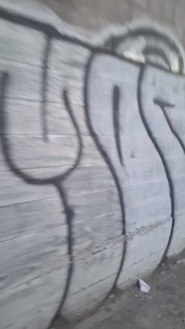 The video shows a person moving rapidly past a concrete wall covered in graffiti. The graffiti consists of stylized lettering in various colors like white, gray, yellow, and blue, and also includes a small cartoonish depiction of a horned character with three eyes and the words "RIP POWIN". The ground near the wall is visible, appearing to be asphalt or concrete. The video is shot from a moving perspective, making the images appear blurry.