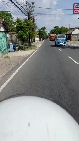 The video begins with a view from a motorcycle, showing a road with traffic. A blue truck with a tank passes by. Further down the road, an orange truck is visible. As the motorcycle continues, it passes a person riding a blue scooter. The person on the scooter is wearing a helmet and a light-colored shirt. The environment appears to be a rural or suburban area with buildings and trees along the roadside. The sky is partly cloudy.
