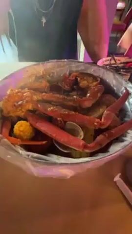 The video begins focused on a metal bowl of seafood, including crab legs, corn on the cob, and clams, inside of a clear plastic bag. A person standing behind the bowl takes a picture of the food with their phone. The camera pulls back and up, revealing the person, wearing a black tank top, seated at the table. They use utensils to eat. The camera pans to the right, showcasing a bar area with purple lighting, chairs, and shelves of bottles. Other customers can be seen in the background.