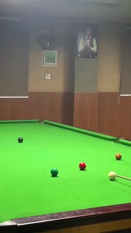 A snooker table with several colored balls is visible. A person is playing snooker, but only their hand and cue are partially visible. The room appears to be a snooker hall, with a poster of a man playing snooker on the wall. The lighting is dim.