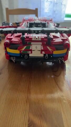 A red LEGO model of a Ferrari race car sits on a wooden table. The car has its doors open, revealing some of the interior mechanisms. The camera slowly pans from the rear of the car to the front, providing a full view of the model. The background is blurred, showing a chair and a window.
