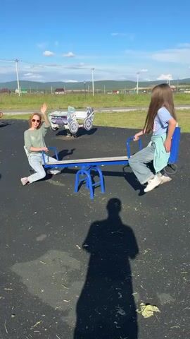 The media file shows two people, an adult woman and a child, on a seesaw in a playground. The playground has a dark, rubberized surface with scattered leaves. In the background, there is a grassy area with a fence and some houses, suggesting a rural or suburban setting. There are also some playground equipment in the background, including what looks like a toy car. The sky is blue with some scattered clouds, and the sun is shining brightly, indicating it is daytime, likely afternoon. The woman is wearing a green long-sleeved shirt, blue jeans, and pink shoes. The child is wearing a light blue t-shirt and blue jeans. Both appear to be enjoying themselves, with smiles on their faces. The woman is on one side of the seesaw, and the child is on the other, with the seesaw in motion. The overall mood is lighthearted and playful. The time of day is likely late afternoon based on the long shadow of the person filming. The location context provided is Mursalimkino, Russia. There is no visible text in the media file.