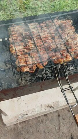 The video shows a close-up, high-angle shot of chicken pieces cooking on a grill over hot coals. The chicken is arranged in a grill basket, and smoke rises from the meat as it cooks. The grill is situated in an outdoor setting, with hints of green foliage and a concrete surface visible in the background. The overall scene depicts a typical barbecue or grilling scenario.