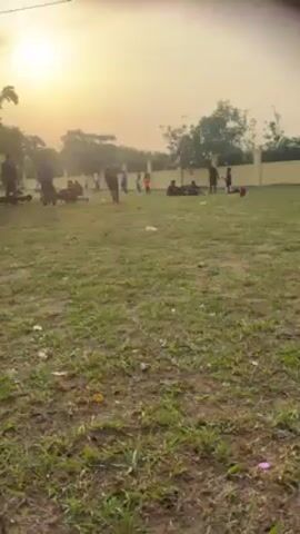 The video shows a grassy field with a group of people, mostly young men, engaged in various activities. Some are doing sit-ups, others are sitting or standing around, and a few are moving about the field. The overall impression is one of group exercise or training. There's a low yellow wall in the background. The sun is visible in the sky, creating a bright but hazy atmosphere. The camera is handheld and provides a ground-level perspective of the scene, and a person's legs can be seen in the foreground.