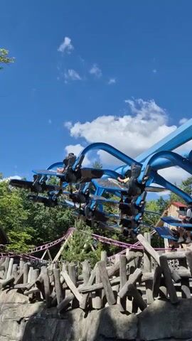 A blue amusement park ride with multiple cars filled with passengers is in motion against a bright blue sky with scattered white clouds. The ride features curved blue arms extending from a central structure, with individual cars suspended from the ends of these arms. The cars are positioned at various angles, with the passengers facing outwards. Below the ride, a purple roller coaster track is visible, weaving through lush green trees. In the foreground, a rustic fence made of rough-hewn logs and stones forms a barrier. The overall scene depicts a sunny day at an amusement park.