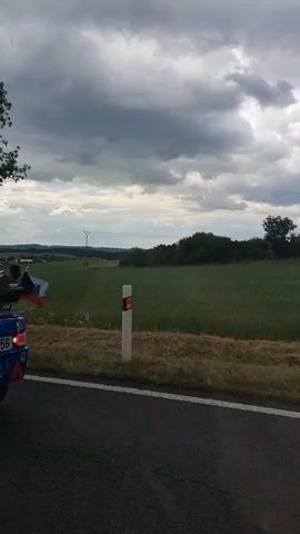 A blue and black motorcycle with a rider wearing a helmet is seen on a road. It is followed by a blue trailer with a black storage box on top. The background is a green field with trees and a cloudy sky. The video seems to be taken from another vehicle moving alongside the motorcycle.