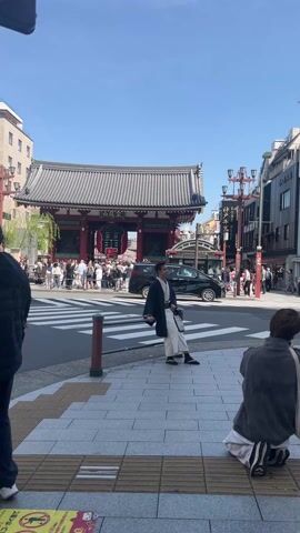The scene is a sunny day in front of Sensō-ji temple in Tokyo. In the foreground, a man wearing traditional Japanese clothing stands on a crosswalk, posing for a photo being taken by a person kneeling on the sidewalk with a camera. Behind them, a crowd of people are milling about near the temple gate. A black van is driving across the crosswalk. The surrounding area includes buildings with traditional and modern architecture, and Japanese street lamps.