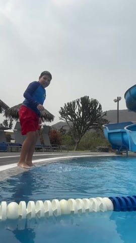 A boy is jumping into a swimming pool. The boy has dark hair and is wearing a blue long-sleeved shirt and red shorts. He is standing on the edge of the pool and smiles at the camera. He then jumps into the pool, creating a big splash. The pool is blue with white and blue lane dividers. In the background, there is a blue water slide, some lounge chairs under thatched umbrellas, a large cactus-like tree, a light pole, and a low mountain range. The sky is overcast.