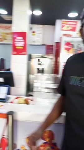 Here is a description of the video:

The video takes place at a fast-food restaurant.  A cashier wearing a red uniform is standing behind the counter, waiting to take orders from customers. Several customers are standing in line, including a woman with braided hair wearing a patterned blue and white shirt, and a man with a black cap and t-shirt. He turns to the camera, and a man with dreadlocks standing nearby turns towards the camera too. Other customers and restaurant details are seen in the background.