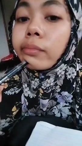 The media file is a close-up, first-person perspective video of a young woman wearing a floral hijab. She is holding a black pen in her right hand, close to her mouth, as if contemplating or about to write. In front of her is a notebook with lined paper, also visible. The woman's facial expression is neutral, with her gaze directed slightly downwards. The background is blurred but appears to be an indoor setting with a wall. The lighting suggests it is daytime. The visible text on the pen reads "UOYOHO". The context of Watampone, Indonesia, provides a geographical setting for this personal moment.