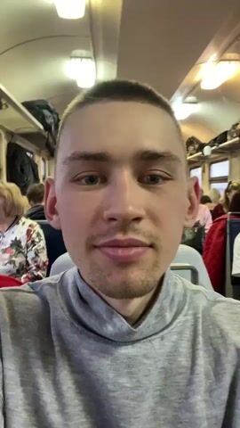 The video is taken from the perspective of a young man with closely cropped brown hair, light skin, and blue eyes. He's wearing a grey turtleneck sweater. He is on a train, as evidenced by the interior of the train car, filled with rows of blue seats and overhead luggage racks. The train is populated with other passengers of varying ages, including women and men, seated and standing. Some passengers are looking at their phones, while others are engaged in conversations. The train car is illuminated by fluorescent lights. There is a visible window with outside scenery. The camera is held relatively steady, showing the man's face as well as the other passengers around him.