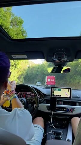 The video shows the interior of a car driving on a road lined with trees and foliage. The driver, with purple hair, holds a bottle of Fanta in their left hand. A navigation system is displayed on the car's infotainment screen, showing a route. A DPD delivery van is visible on the road ahead. The car has a sunroof, which is open, revealing a clear blue sky. The camera is positioned from the passenger's seat, looking forward.