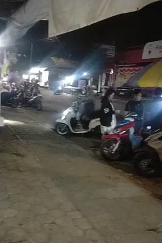 The video shows a nighttime scene with several motorcycles parked.  A person is seated on one of the motorcycles, wearing dark clothing. Another person is standing next to the motorcycle, also in dark clothing. There is a multi-colored umbrella over the motorcycles.  Lights are visible, suggesting this is an outdoor area, possibly a street or market. The overall environment appears to be somewhat cluttered with the parked motorcycles and possibly merchandise or structures in the background.