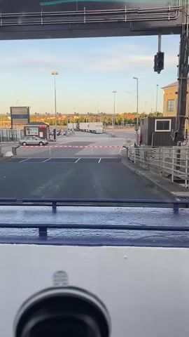 The video begins with a view from a ferry as it departs a terminal. A white ferry railing is visible in the foreground. Beyond the railing, a paved area with white lane markings is seen, leading away from the ferry. Several vehicles, including cars and trucks, are parked and driving in the distance. The sky is blue with some clouds, and the sun appears to be setting, casting a warm light. A traffic light is visible on a pole. The ferry's ramp is down, and it appears to be moving slowly away from the terminal. The video is shot from a slightly elevated perspective, looking forward and slightly downwards.