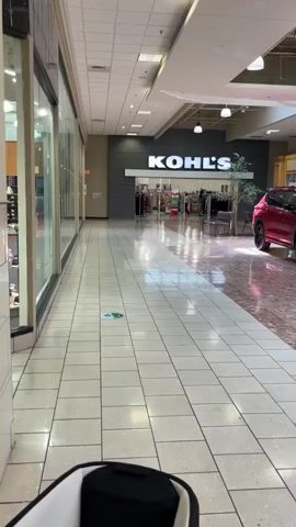 The video shows a view from a shopping cart in a mall hallway. On the left, there are store windows with merchandise. Straight ahead, the hallway leads to a Kohl's department store, which has its logo prominently displayed. To the right of Kohl's, a red SUV is parked in the mall. The floor of the hallway is tiled, with a section of reddish-brown patterned tiles in front of Kohl's. There is a green and white floor sticker visible in the hallway. The lighting suggests it is daytime, and the mall appears to be sparsely populated.